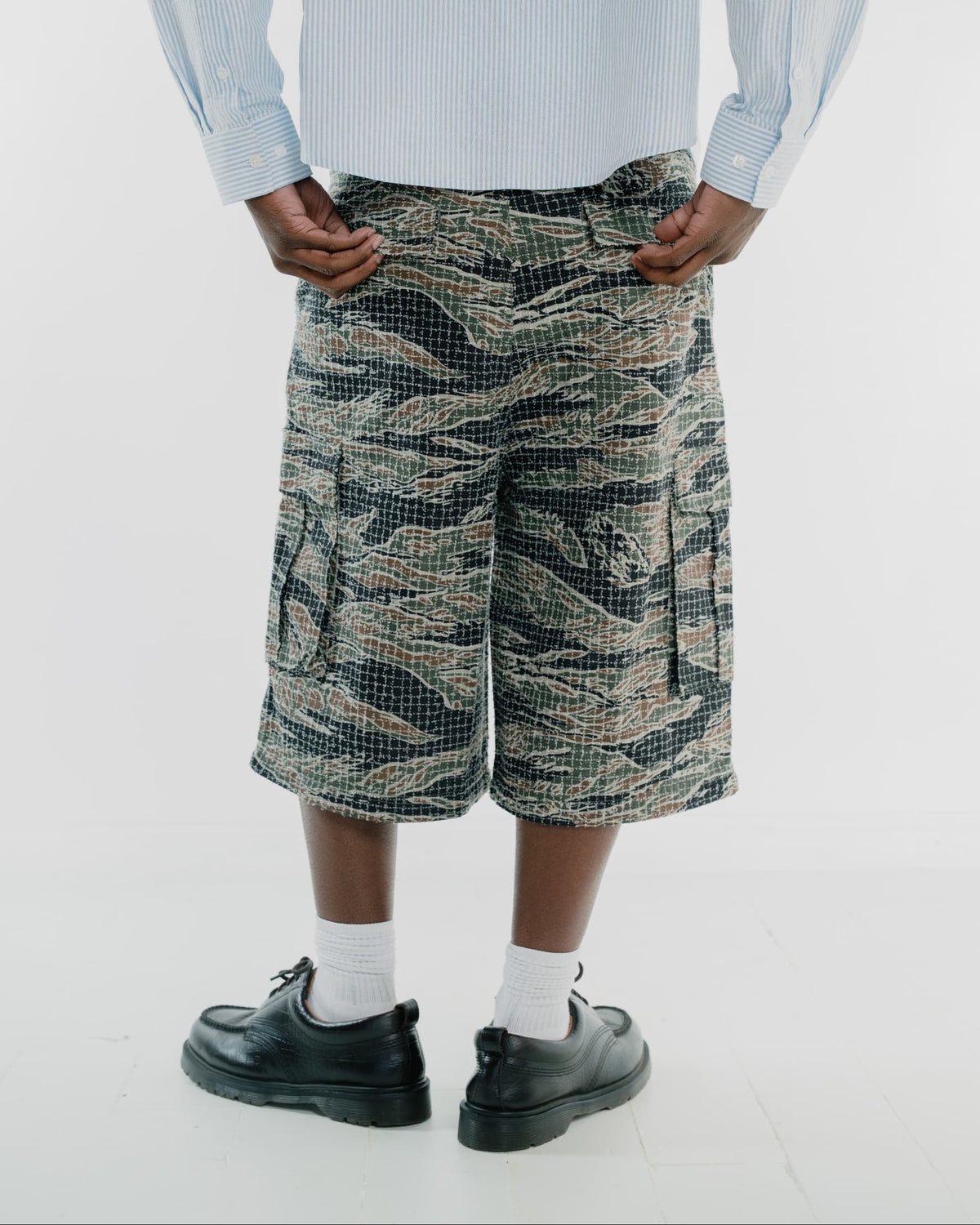 A person shown from the waist down wearing tiger stripe camouflage cargo shorts, a light blue striped shirt, white socks, and black leather shoes against a plain white studio background.