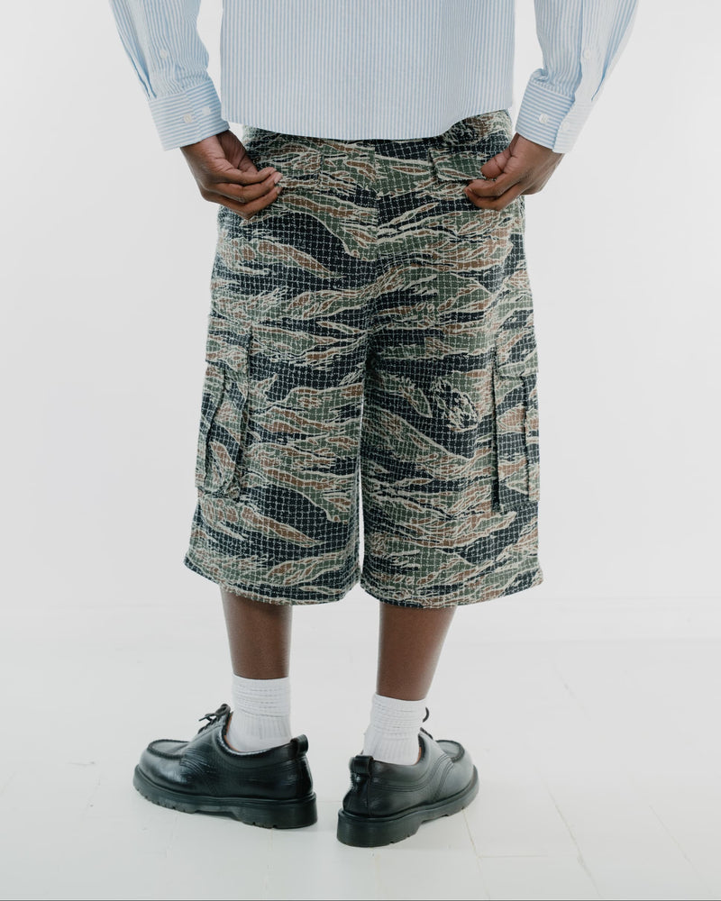 A person shown from the waist down wearing tiger stripe camouflage cargo shorts, a light blue striped shirt, white socks, and black leather shoes against a plain white studio background.