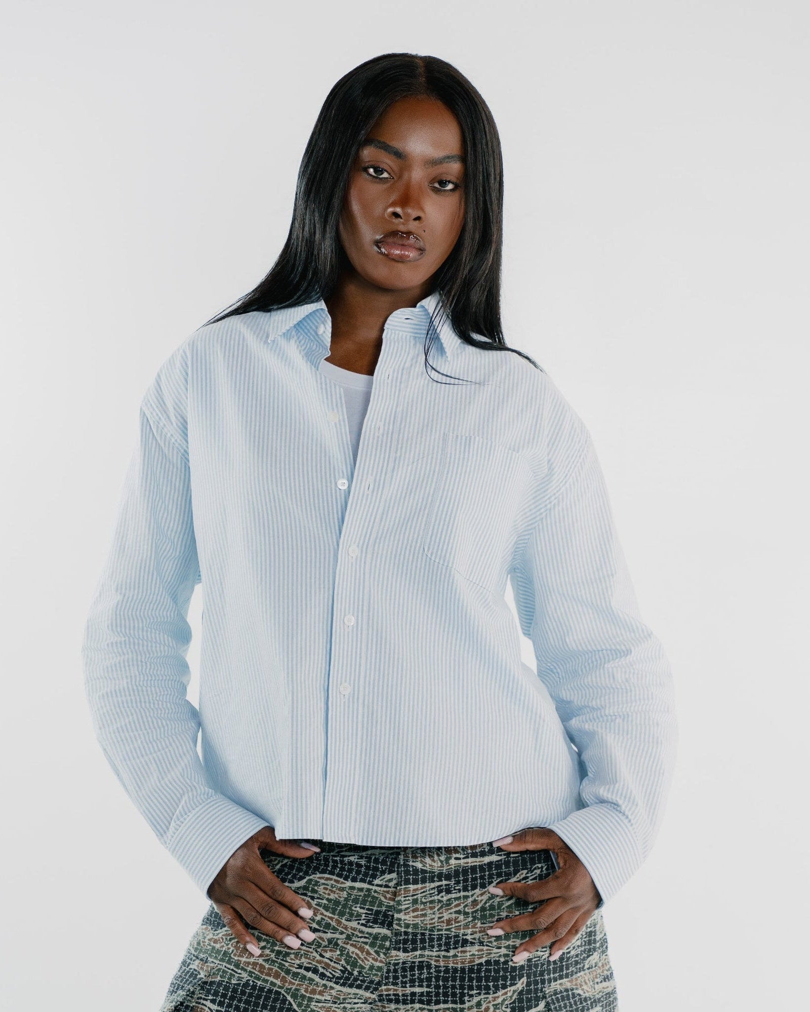 A model wearing a light blue and white vertically striped button-down shirt tucked into green and brown tiger-print cargo pants, shot against a plain white studio background.