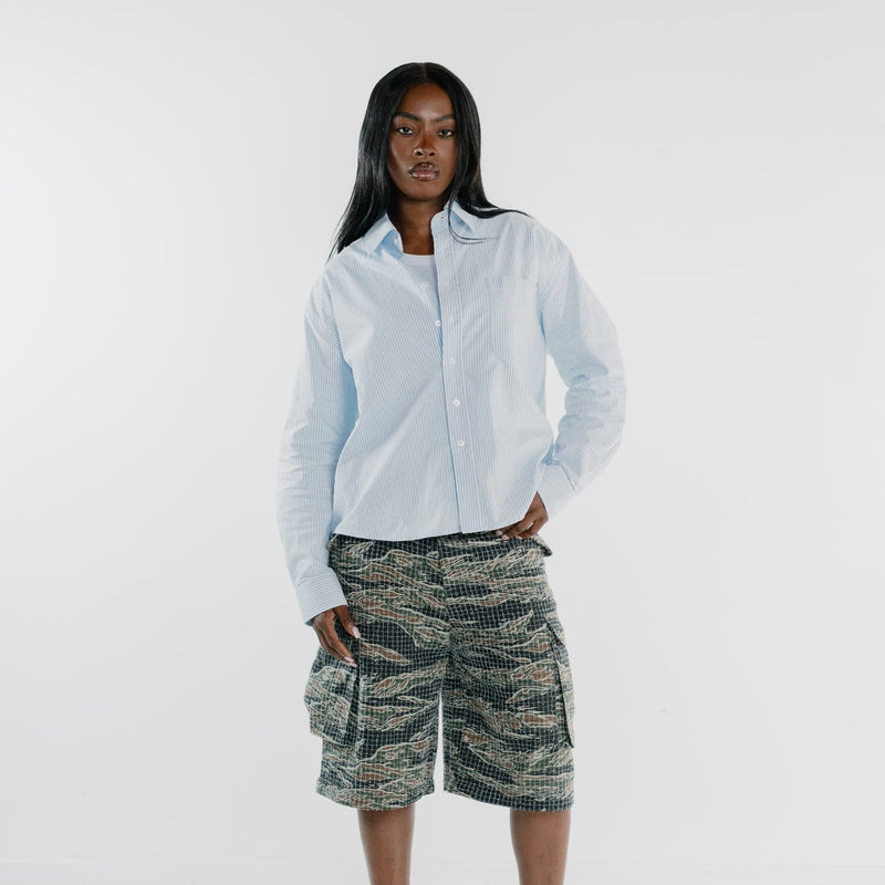 A model stands against a white studio background wearing a light blue long-sleeve button-up shirt, patterned cargo shorts in green, beige, and black, and black slide sandals.
