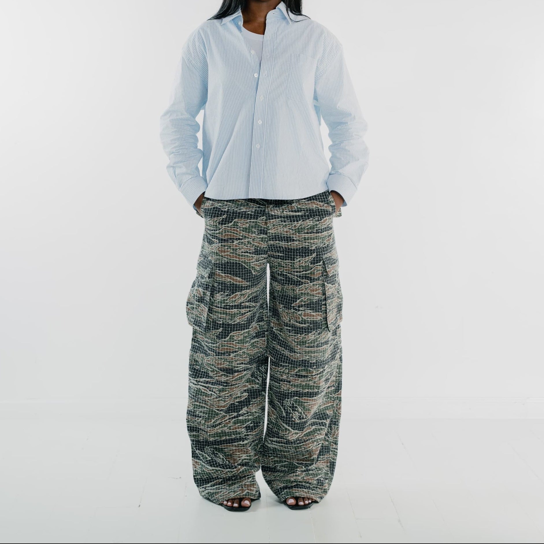 A model stands against a light gray studio background wearing a light blue button-up shirt and wide-leg cargo pants with a green, black, and beige tiger-stripe camouflage pattern.