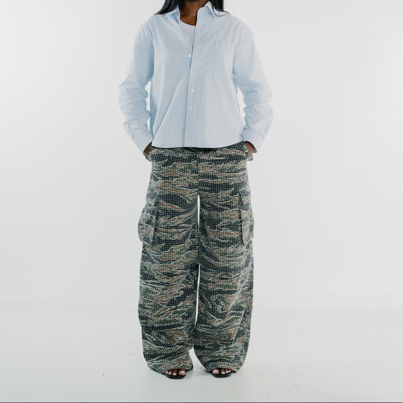 A model stands against a light gray studio background wearing a light blue button-up shirt and wide-leg cargo pants with a green, black, and beige tiger-stripe camouflage pattern.