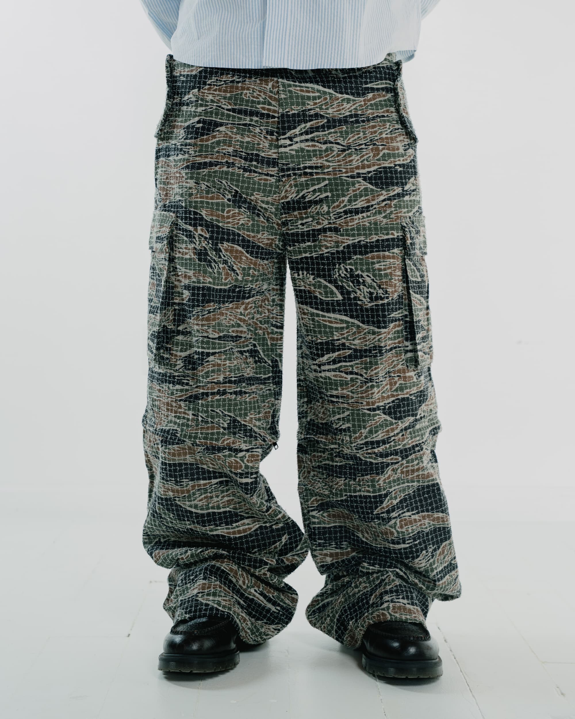 A person wearing baggy camouflage cargo pants with a tiger-stripe style pattern in green, brown, black, and beige, styled with a light blue striped shirt and black shoes against a plain white background.