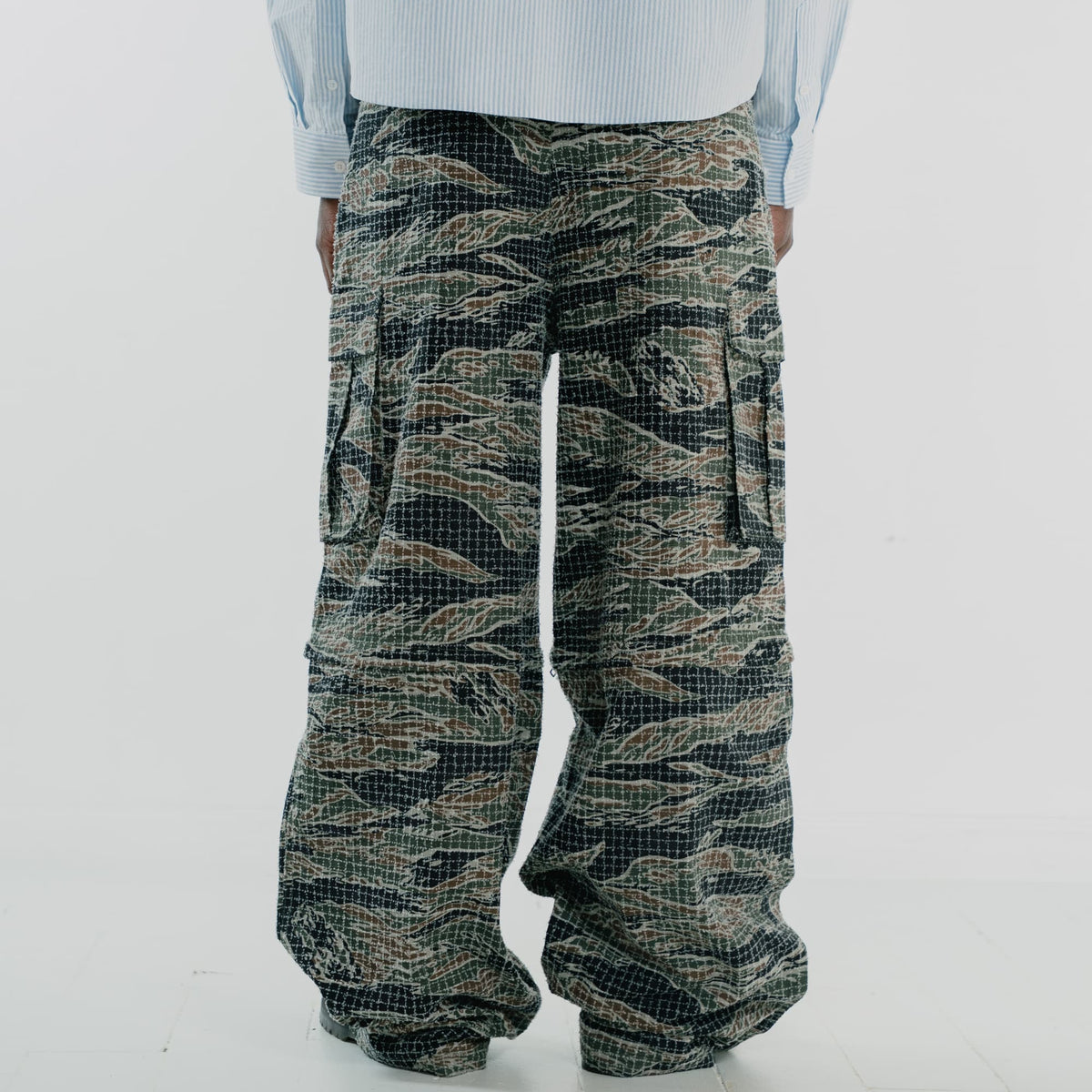 A person shown from the back wearing very wide-leg cargo pants with a tigerstripe camouflage pattern in olive green, black, and tan, paired with a light blue striped shirt against a white studio background.