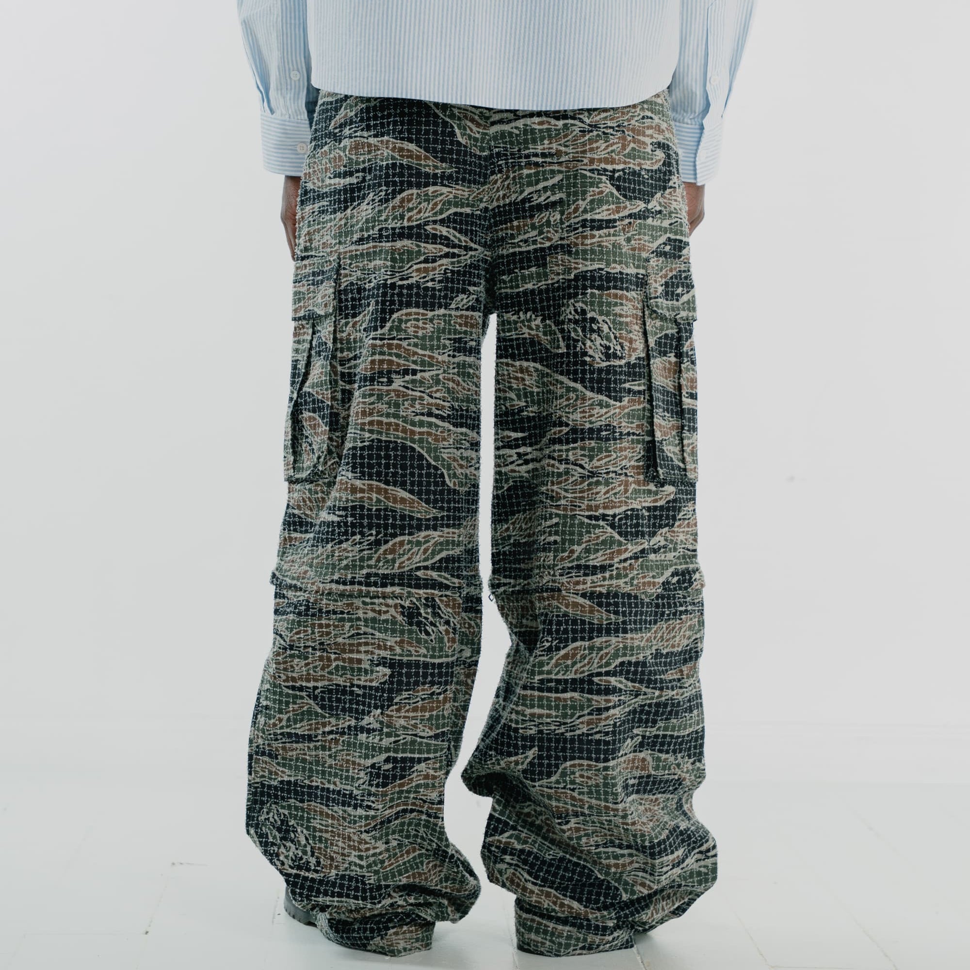 A person shown from the back wearing very wide-leg cargo pants with a tigerstripe camouflage pattern in olive green, black, and tan, paired with a light blue striped shirt against a white studio background.