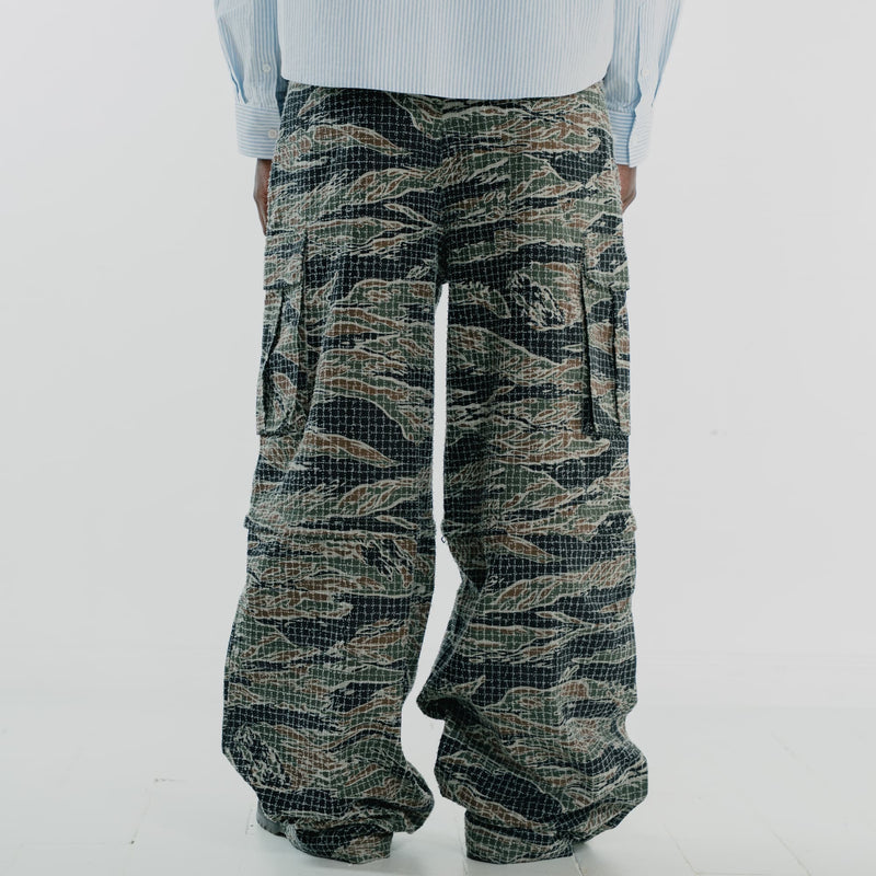 A person shown from the back wearing very wide-leg cargo pants with a tigerstripe camouflage pattern in olive green, black, and tan, paired with a light blue striped shirt against a white studio background.