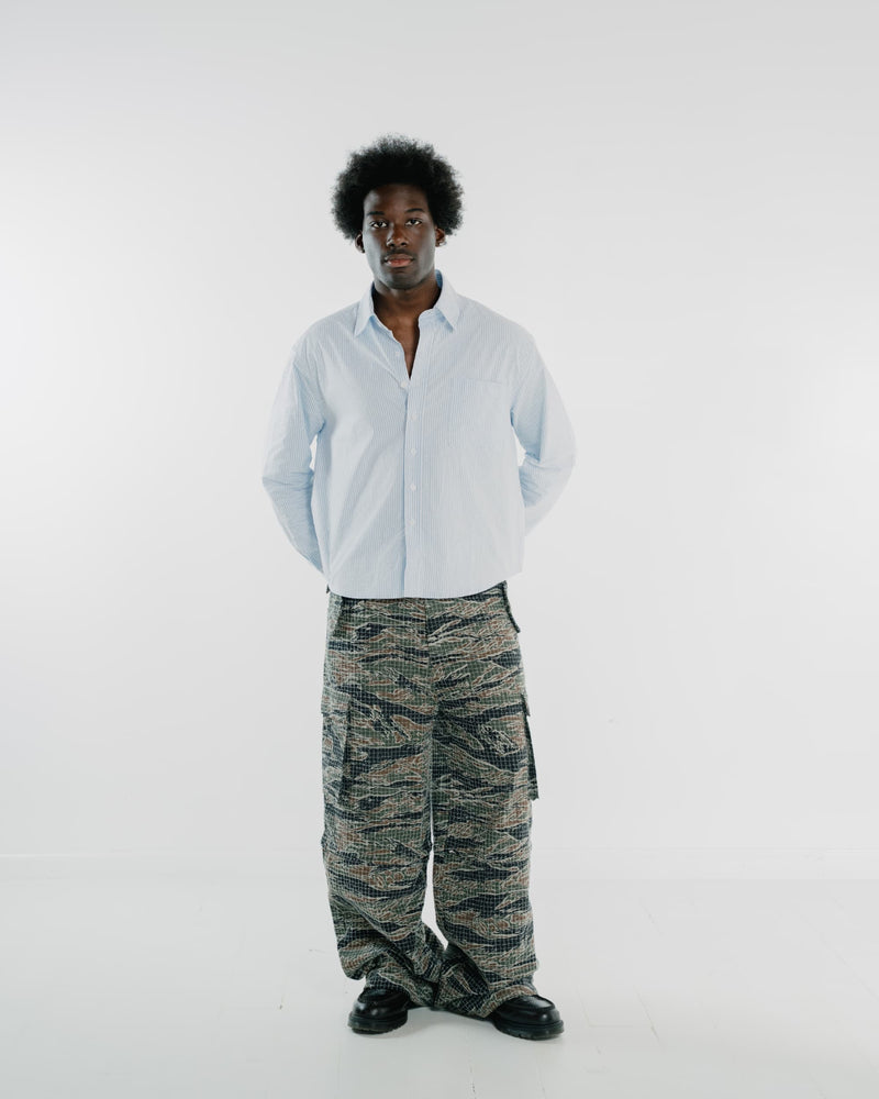 A man stands against a plain light gray studio background wearing a light blue button-up shirt, loose tiger-stripe camouflage cargo pants, and black shoes.