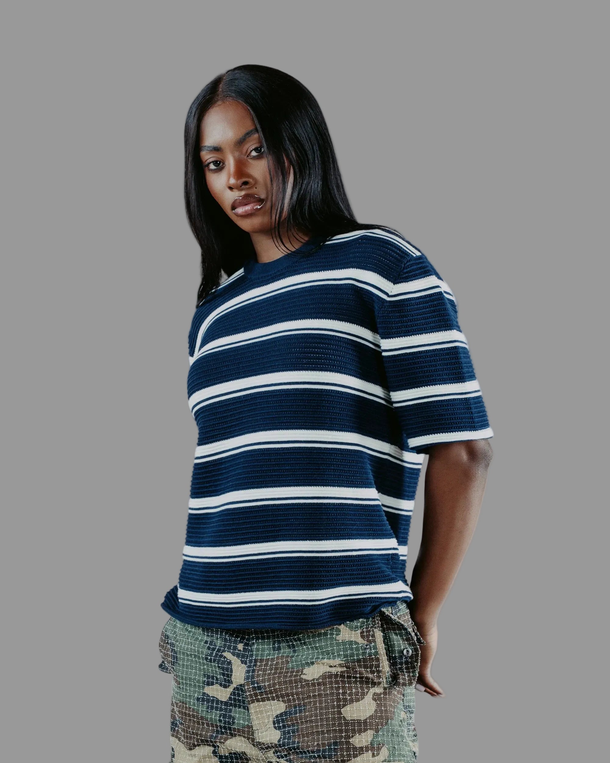A woman wearing a navy blue and white horizontally striped short-sleeve knit top, paired with camouflage shorts, standing against a textured gray wall.
