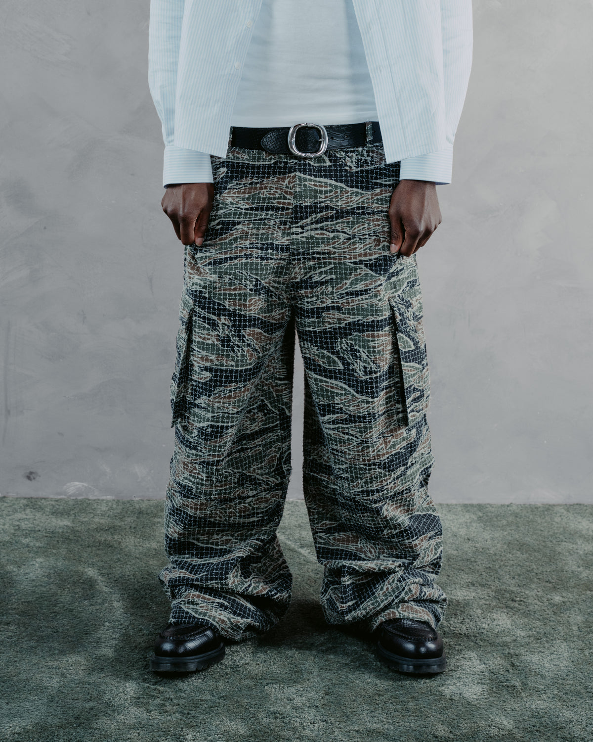 A person wearing oversized cargo pants with a tiger-stripe camouflage pattern in black, gray, and beige, styled with a white shirt, light blue jacket, and black belt.