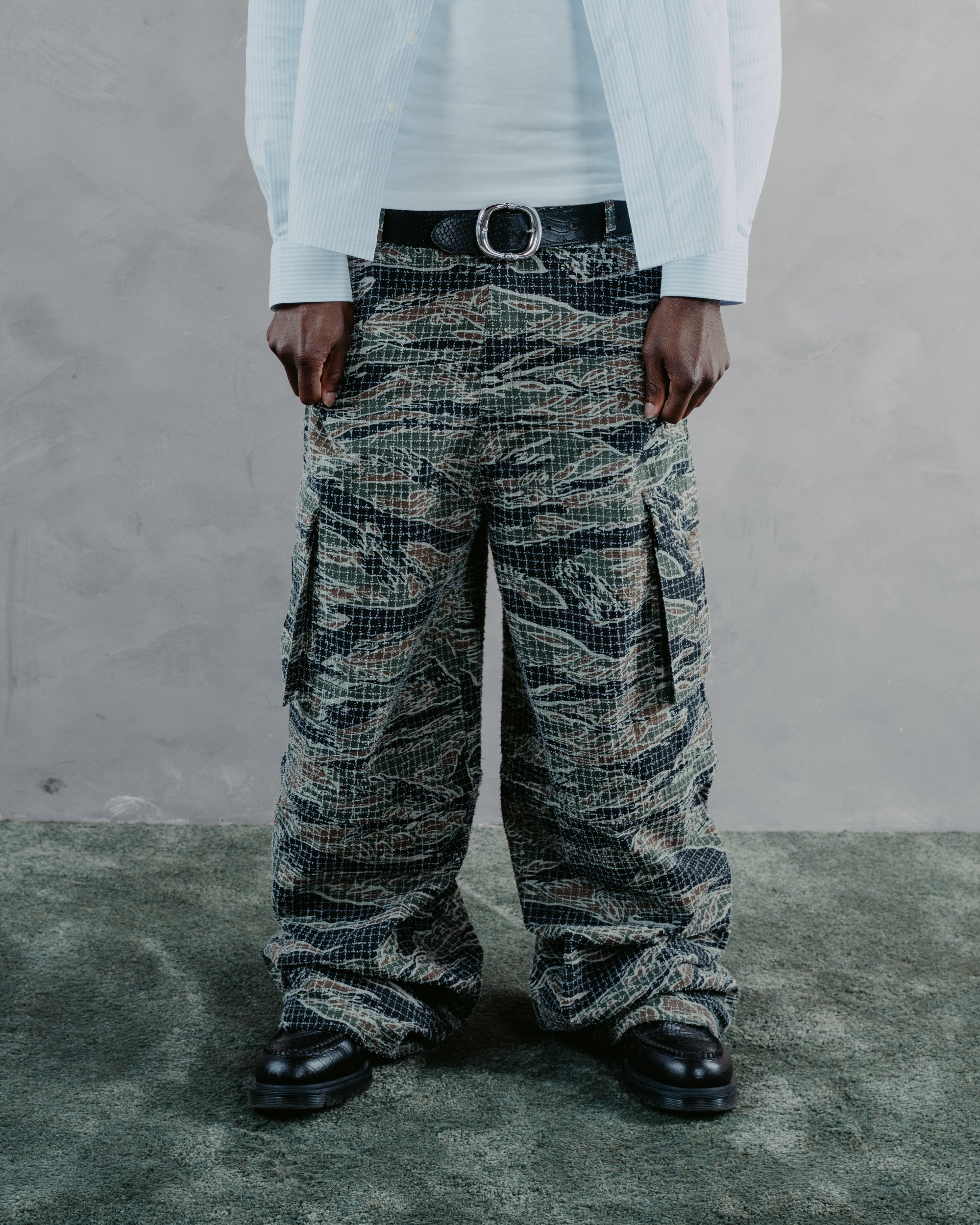 A person wearing oversized cargo pants with a tiger-stripe camouflage pattern in black, gray, and beige, styled with a white shirt, light blue jacket, and black belt.
