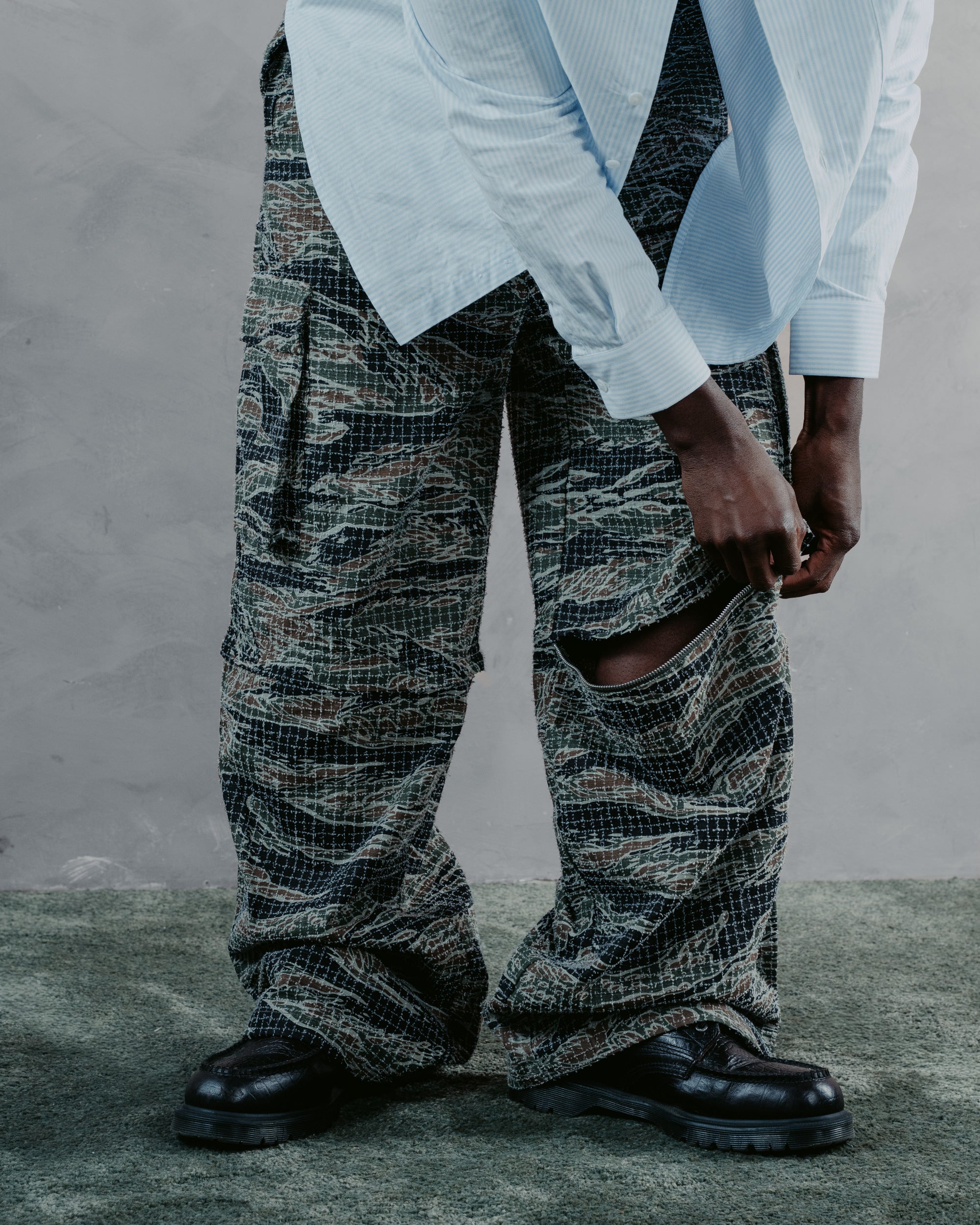A person wearing very wide-leg camouflage pants with a large ripped detail on the leg, styled with a light blue striped shirt and black leather boots against a textured gray wall.