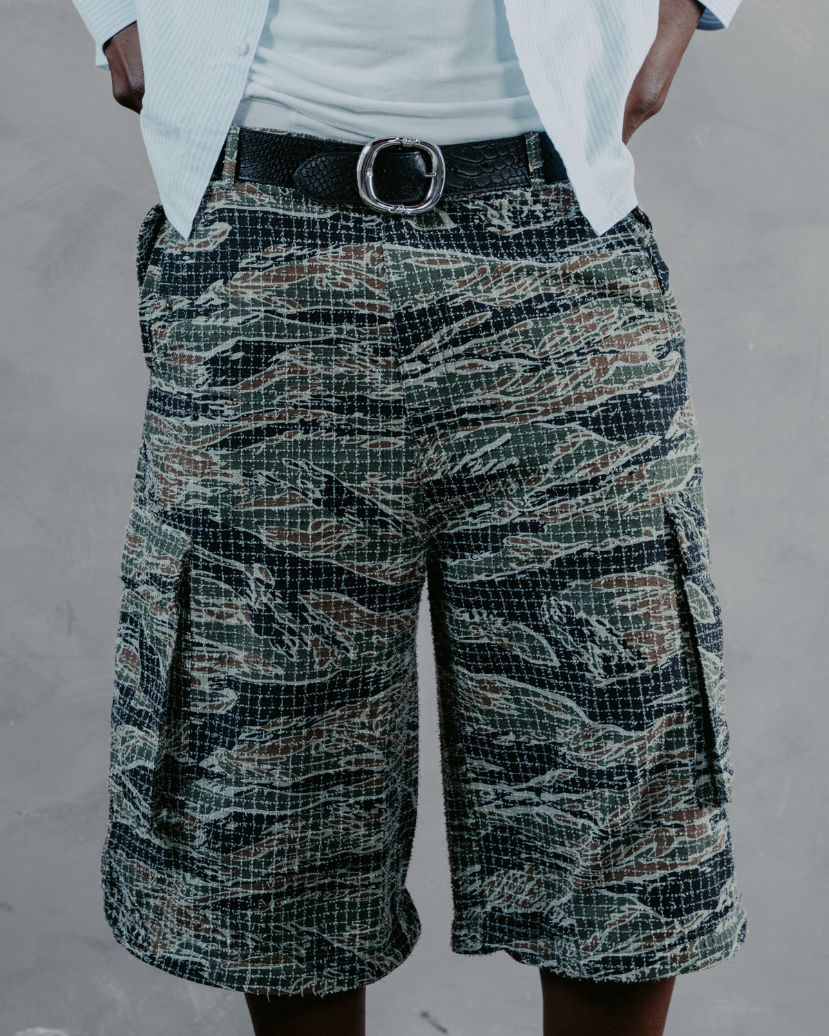 A person wearing tiger-stripe camouflage cargo shorts with a black belt and white shirt, shown from the waist down against a gray textured wall.