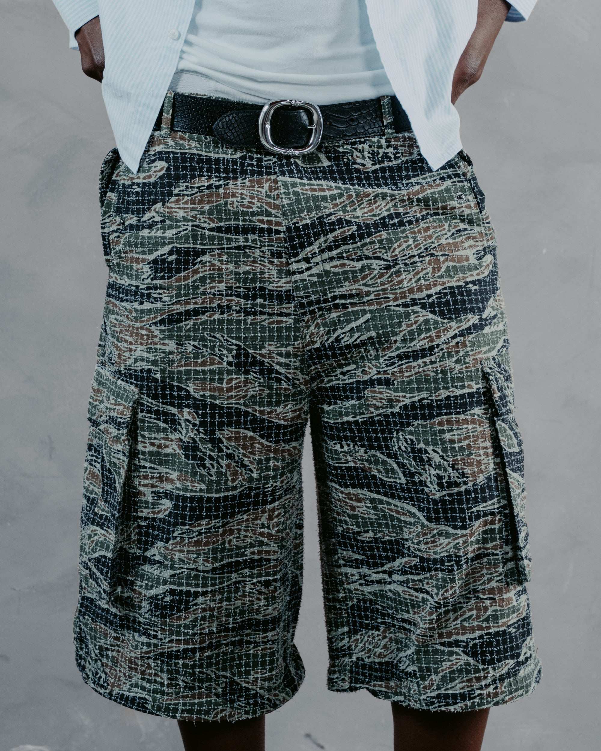 A person wearing tiger-stripe camouflage cargo shorts with a black belt and white shirt, shown from the waist down against a gray textured wall.