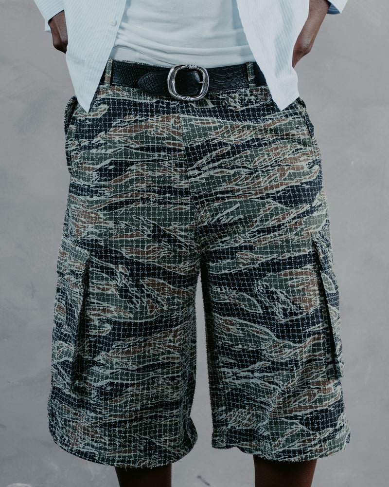 A person wearing tiger-stripe camouflage cargo shorts with a black belt and white shirt, shown from the waist down against a gray textured wall.