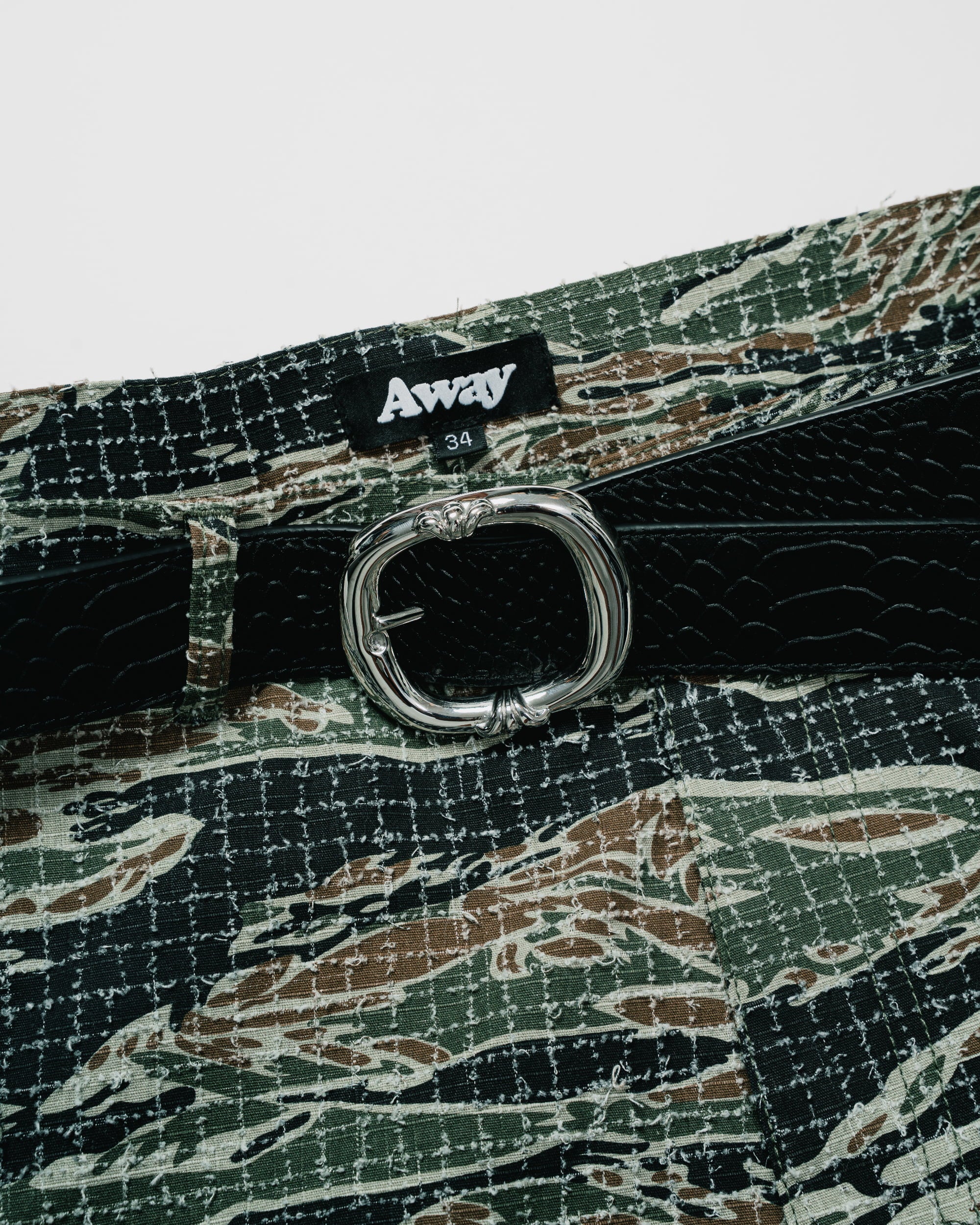 Close-up of a belt with a silver-toned oval buckle, black textured strap, and a green, brown, and beige camouflage patterned fabric body. A black brand label reading 'Away' is visible at the waistband.