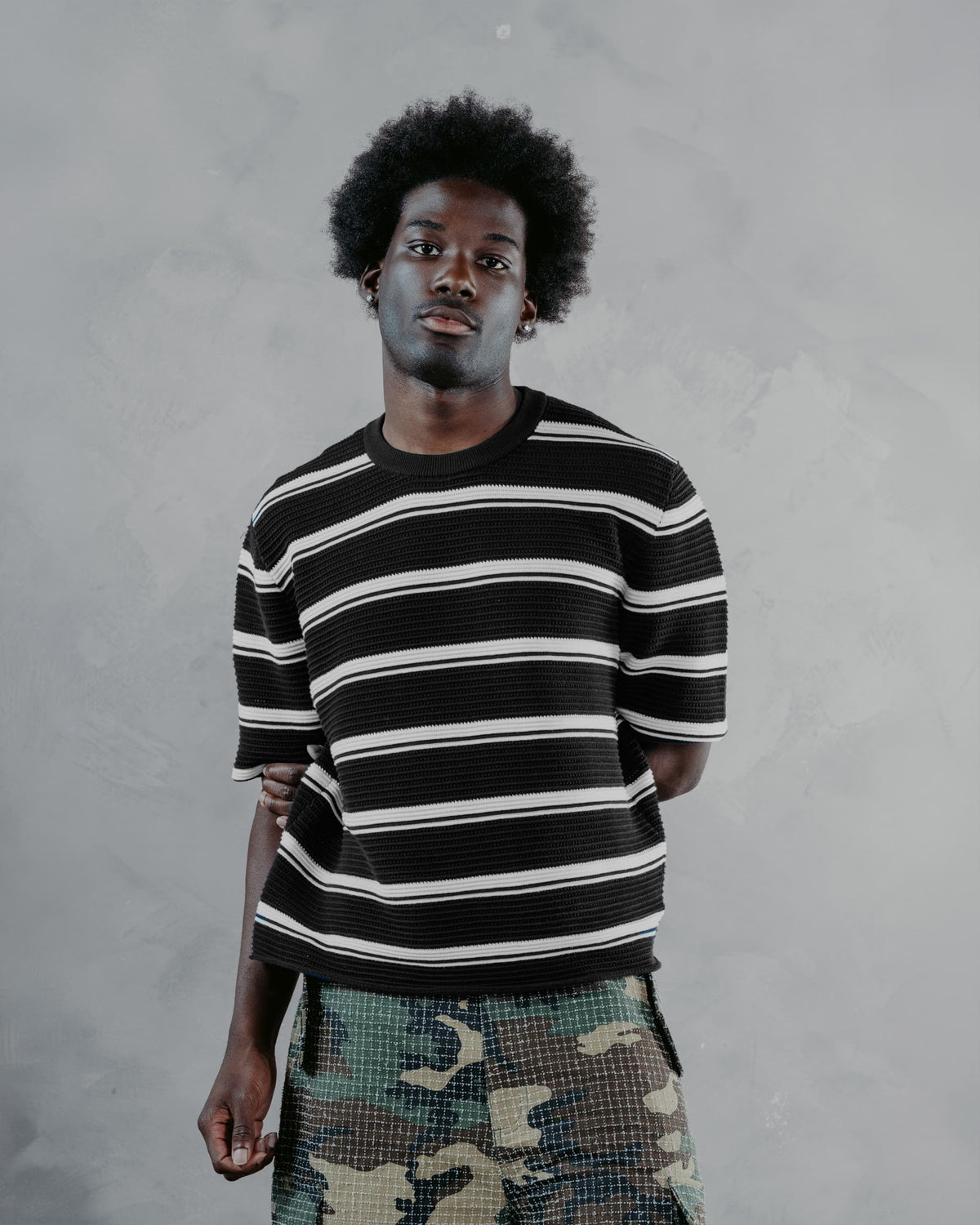 A man with an afro hairstyle wearing a black and white horizontally striped short-sleeve knit top and camouflage pants, standing against a textured gray studio background.