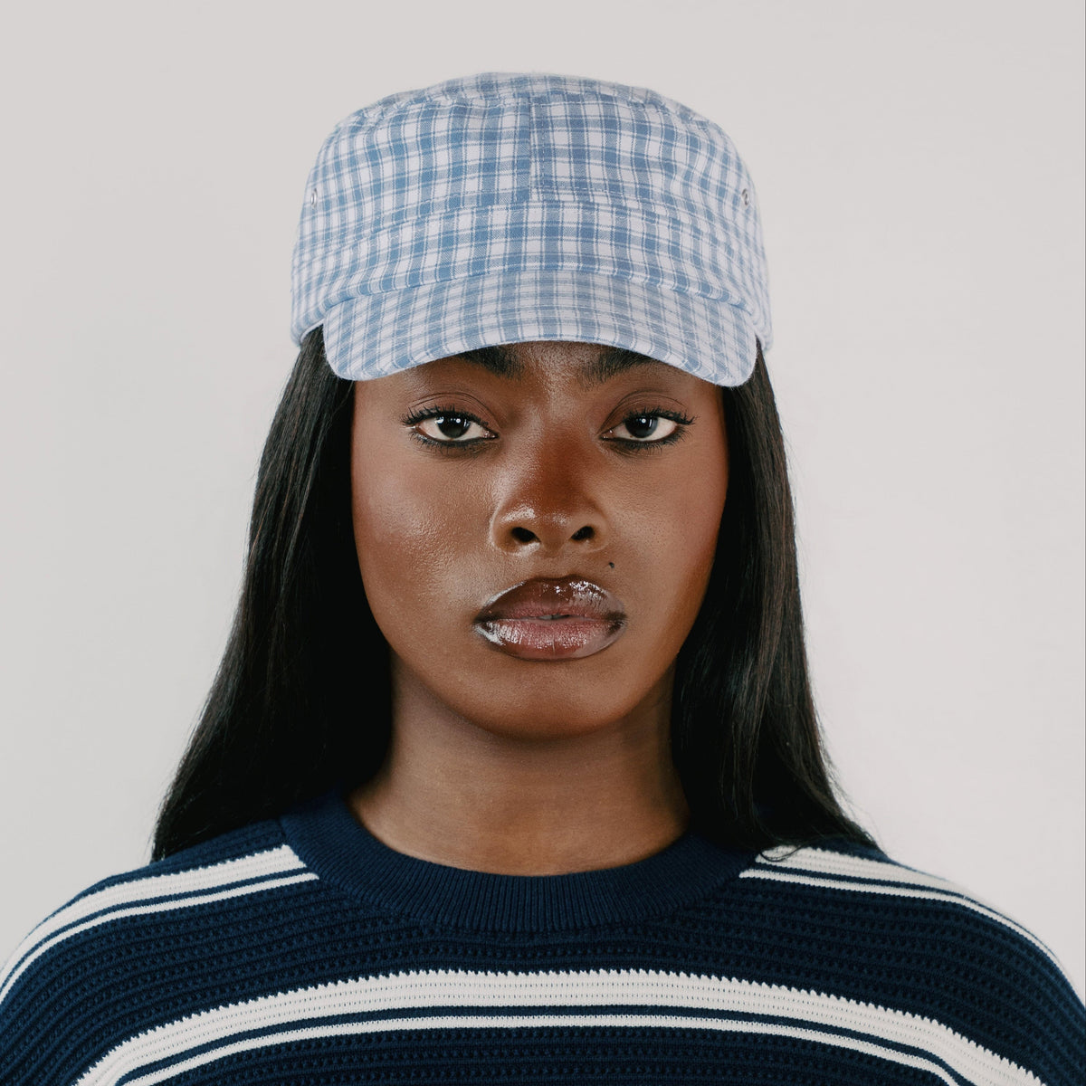 A model wearing a light blue and white gingham checkered cap, paired with a navy blue and white striped knit sweater, against a solid light gray background.