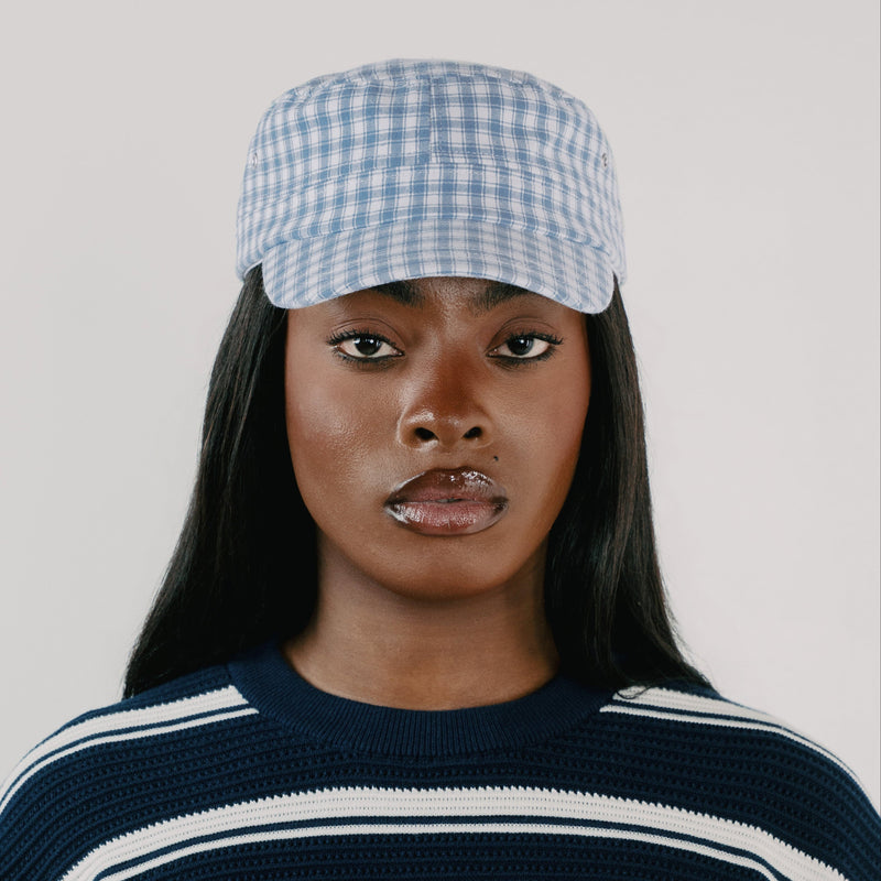 A model wearing a light blue and white gingham checkered cap, paired with a navy blue and white striped knit sweater, against a solid light gray background.